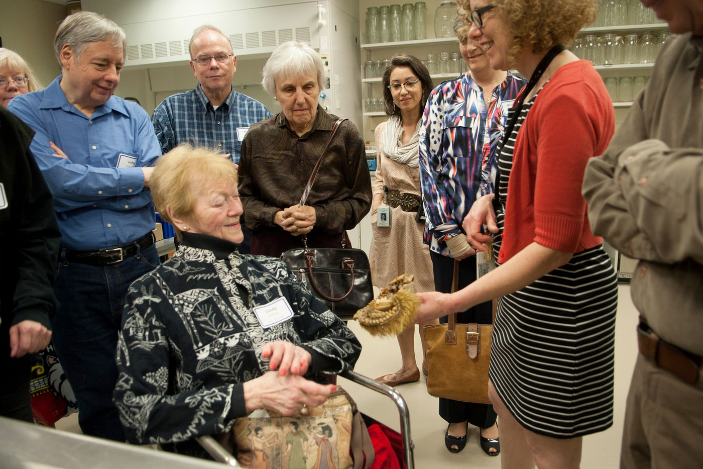 Media for Group Visits An older woman in a wheelchair smiles as she looks at a specimen held by a staff member. Other adults in the group stand around the pair, looking intently at the specimen. Clear jars are on shelves in the background.