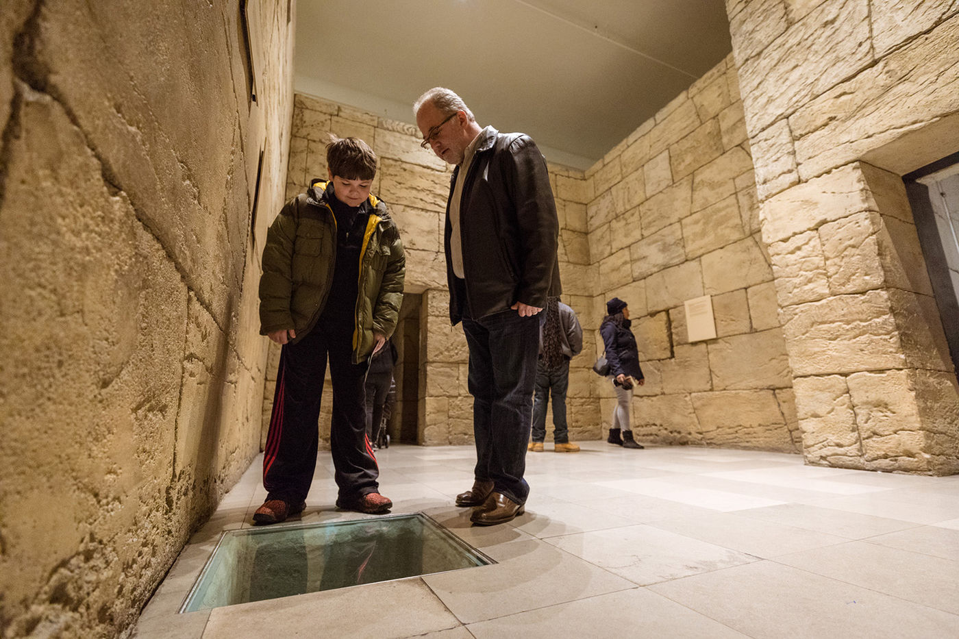 Standing in the ancient Egyptian tomb in the Inside Ancient Egypt exhibition, an adult and child look down at a glass-covered opening in the floor.