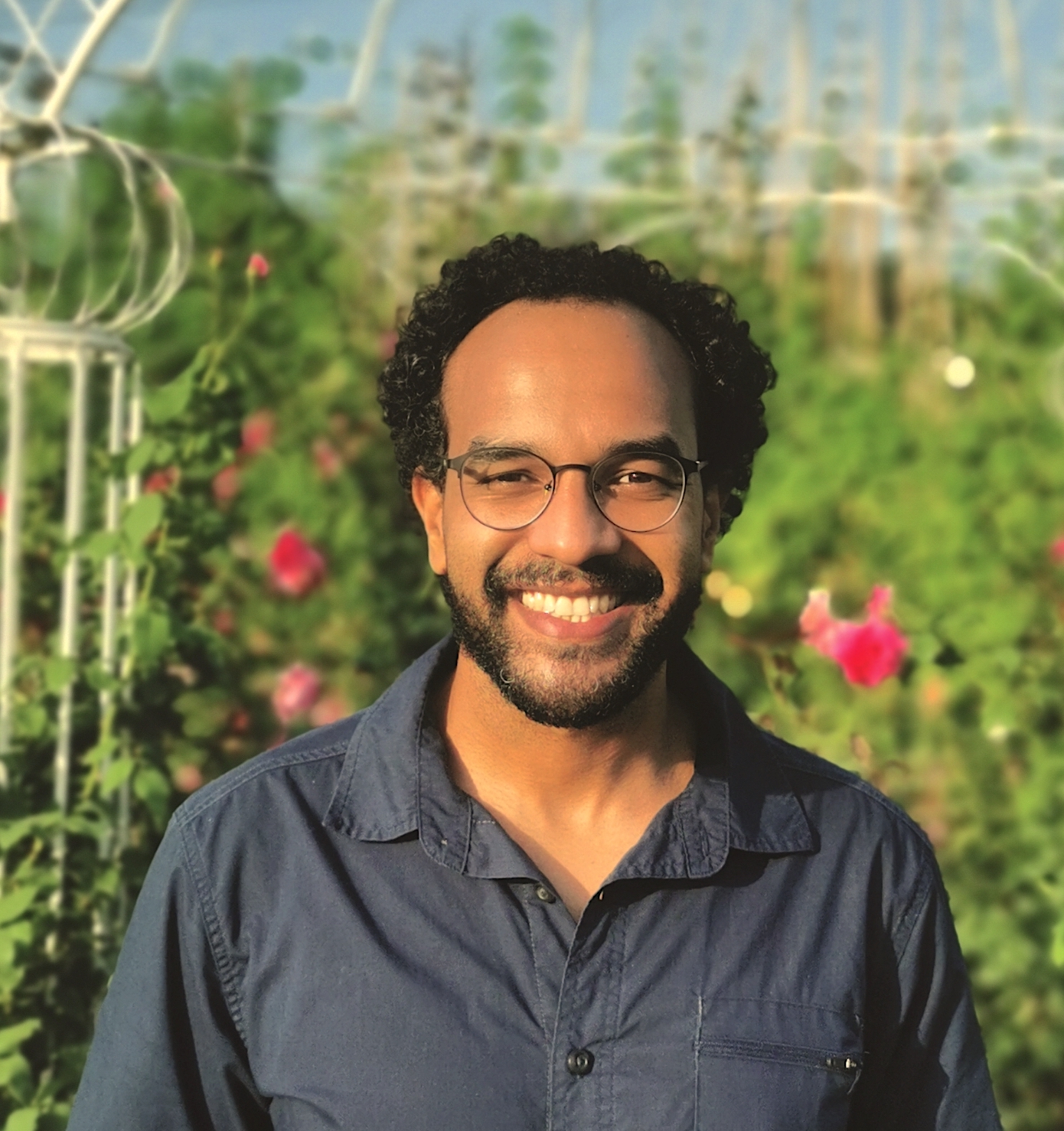 A smiling man with black curly hair and glasses stands in front of a garden.