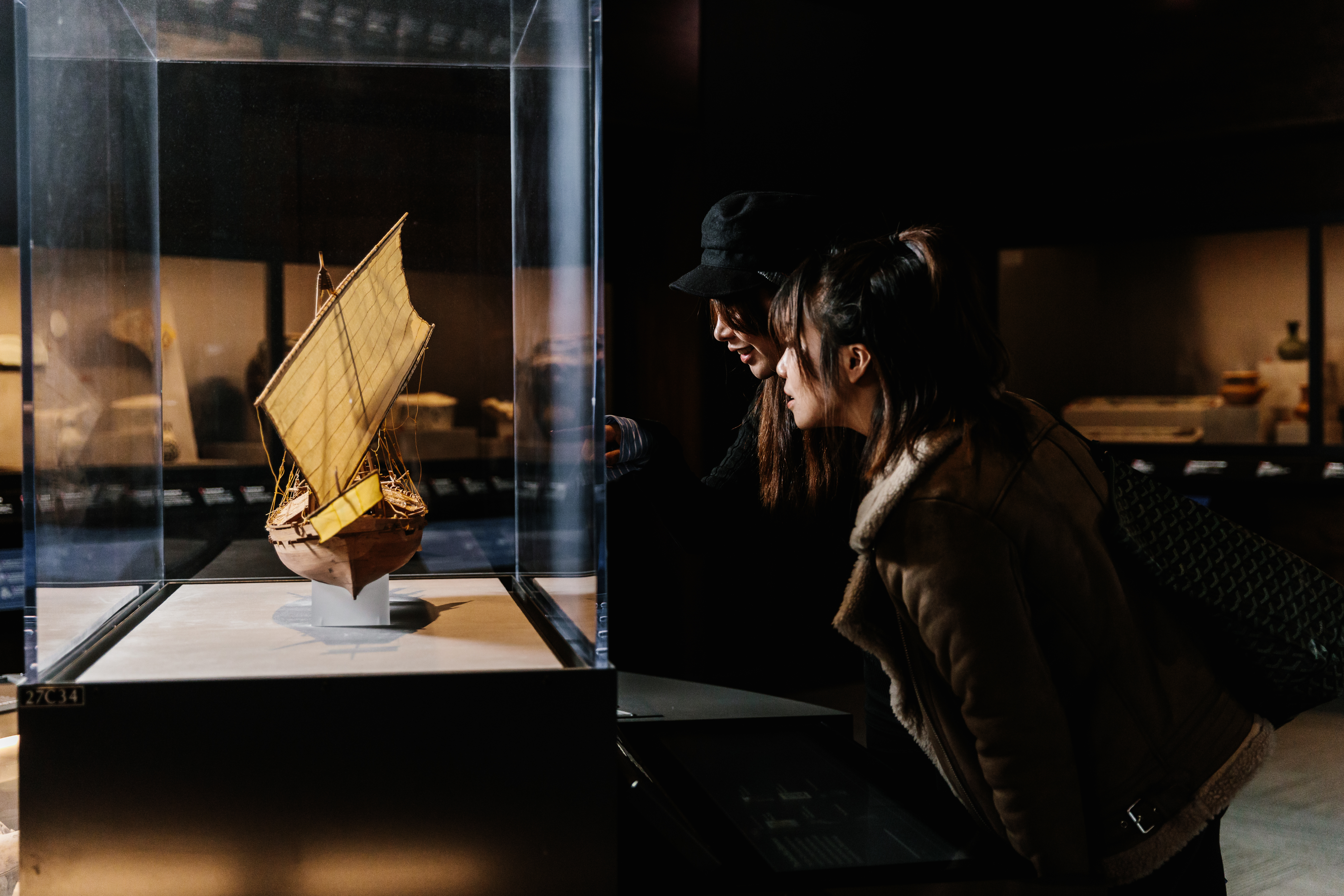 Two museum visitors peer into an exhibition case that features a replica sailing ship.