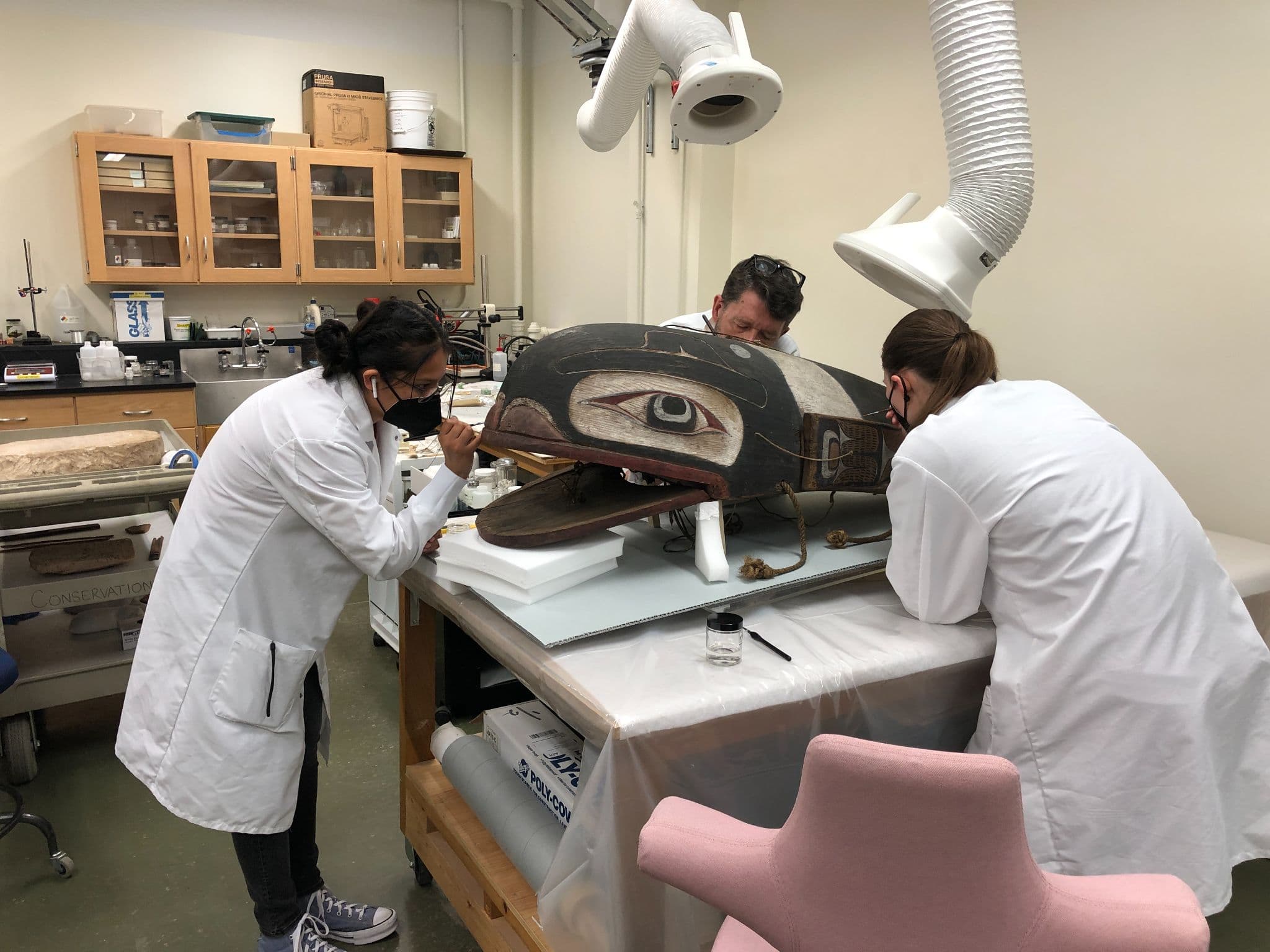Three people dressed in white lab coats lean over a large, carved wooden object on a workbench. They are consolidating the object with small hand tools