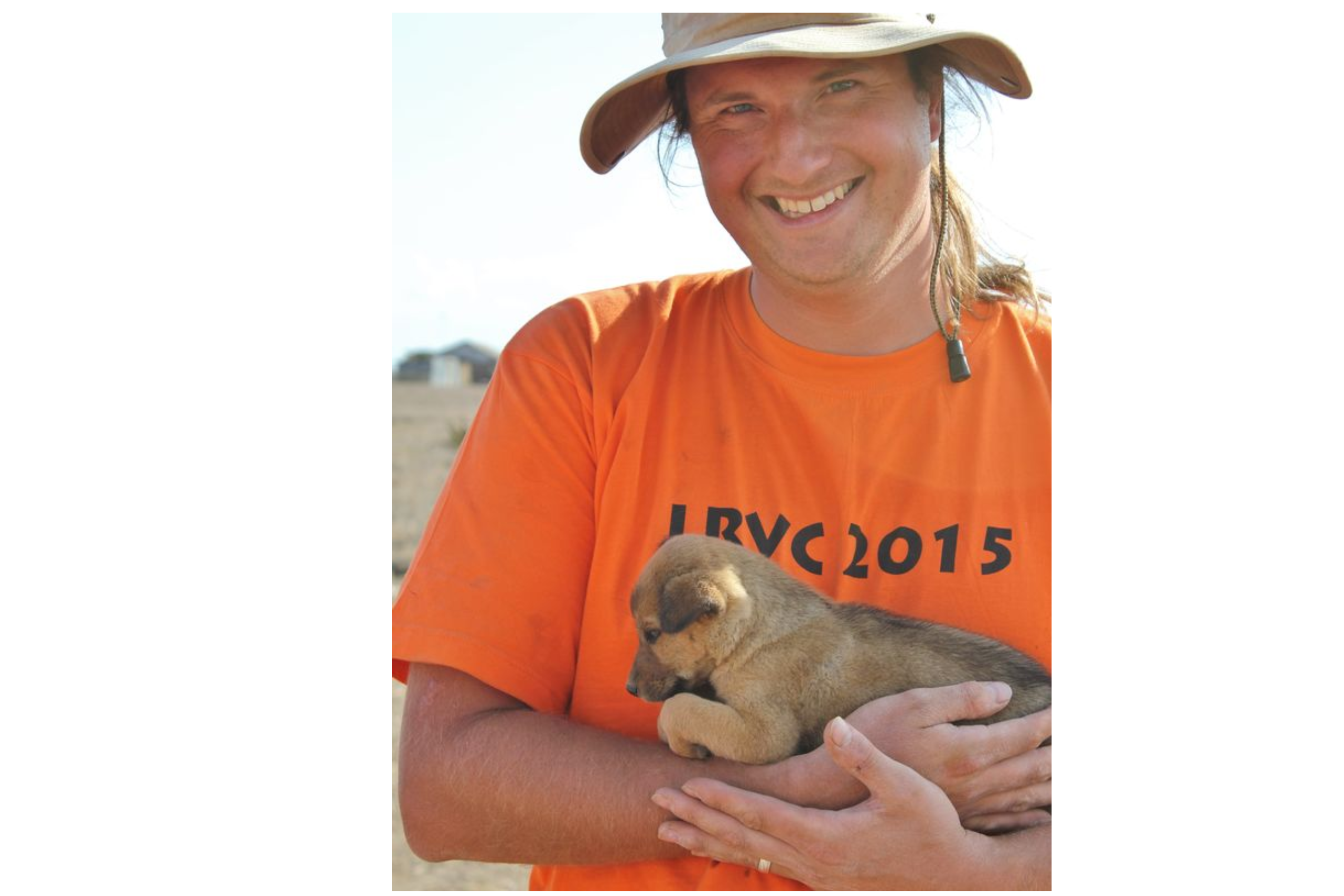 A smiling man in an orange shirt holds a small brown puppy