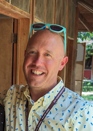 A man, smiling and wearing a button down shirt. His sunglasses are perched on top of his head.