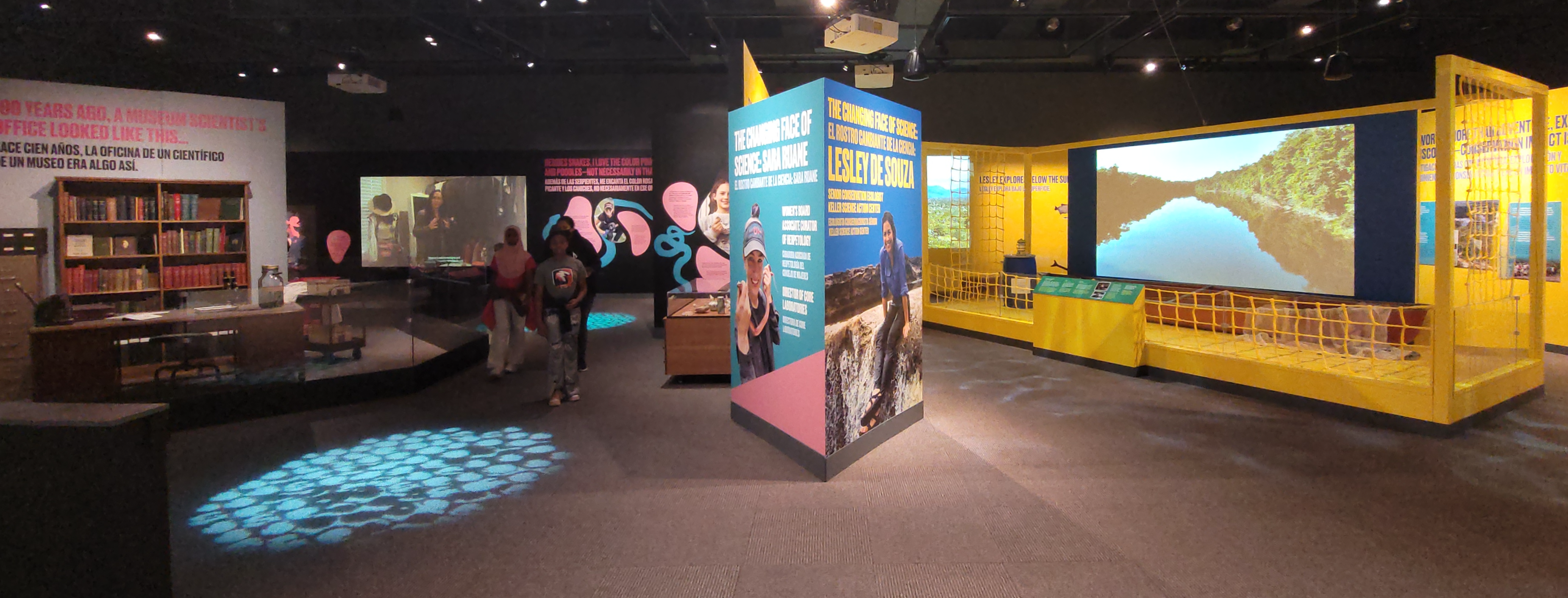 Museum exhibitions featuring two scientists. On the left is a desk and bookshelves, with multimedia panels. On the right, a large video screen and a canoe.